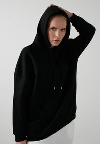 Čierna oversized mikina s kapucňou na šnúrku, klokánkovou kapsou a rebrovanými manžetami. Vyrobená z mäkkej tkaniny s hladkou textúrou.