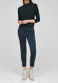 Pull en maille vert foncé avec un col montant et des détails décoratifs en perles, associé à un pantalon tailleur bleu marine et des bottines beige clair.