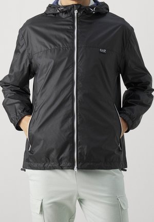 Mann trägt eine schwarze, mit Reißverschluss versehene Kapuzenjacke mit elastischen Bündchen und Händen in den Reißverschlusstaschen, kombiniert mit hellen Cargo-Hosen.