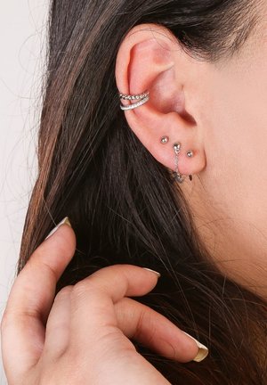 Zilveren oorjuwelen met meerdere studs, cuffs en kettingen met ingewikkelde details, gedragen aan een menselijke oor met lang donker haar.