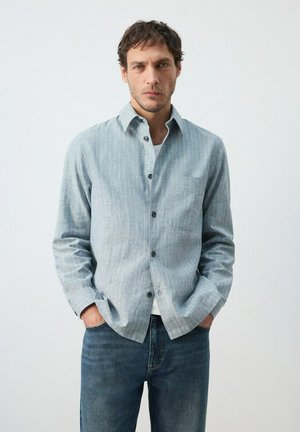 Hombre con cabello corto y oscuro que lleva una camisa gris clara con rayas y botones sobre una camiseta blanca, jeans azules, de pie con las manos en los bolsillos.