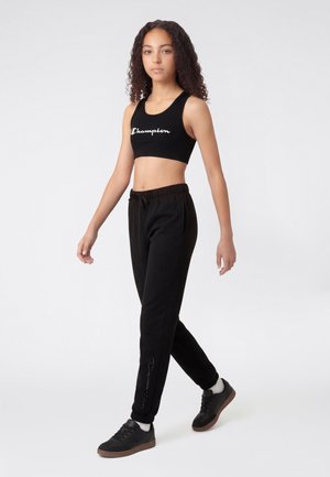 Junge Frau, die in einem schwarzen Crop-Top mit „Champion“-Logo, schwarzen Jogginghosen, weißen Socken und schwarzen Sneakers vor einfarbigem Hintergrund läuft.