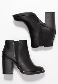 Botas de tobillo negras hechas de cuero sintético, con un tacón grueso, paneles laterales elásticos y un acento dorado a lo largo de la suela.
