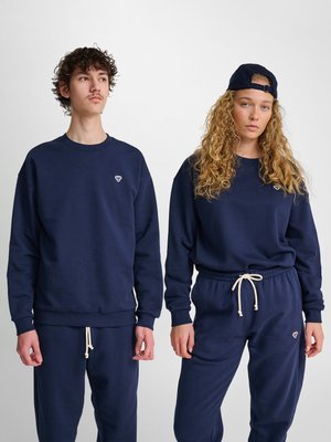 Deux jeunes adultes portant des sweats à capuche et pantalons de survêtement bleu marine avec de petits logos, l’un portant une casquette bleu marine à l’envers, sur un fond uni.