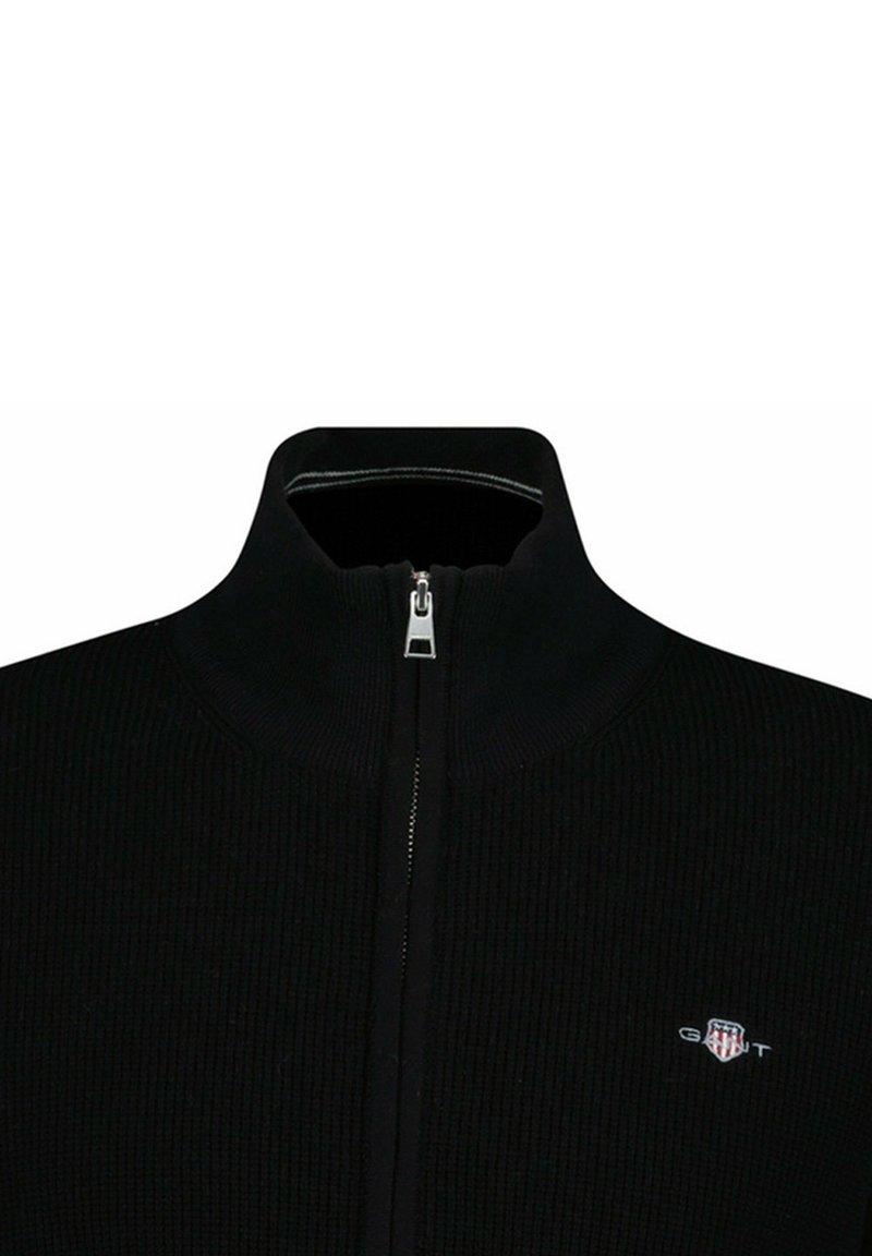 Suéter negro con cremallera, textura acanalada, cuello alto y cremallera plateada. Presenta un pequeño logotipo en el lado izquierdo del pecho.