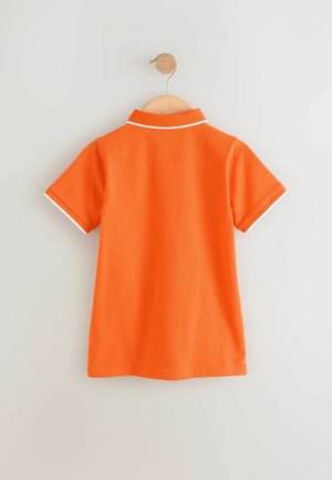 Maglietta polo arancione appesa a un appendiabiti in legno, con colletto e bordi delle maniche bianchi. Tessuto liscio, maniche corte e design classico.