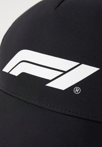 Czarna czapka z wyraźnym białym logo Formuły 1. Materiał jest gładki, ma ustrukturyzowany kształt i przeszywaną szew na daszku.