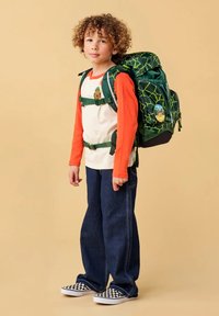 Sac à dos à motif vert avec des bretelles ajustables, porté sur une chemise blanche avec des manches longues orange, associé à un jean bleu foncé et des chaussures noir et blanc.