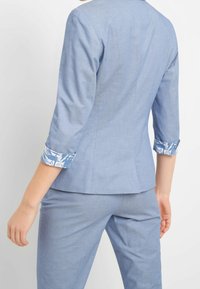 Svetle modré sako na míru s tříčtvrtečními rukávy, zdobené květinovým vzorem v modré a bílé na manžetách. Hladká, strukturovaná tkanina.