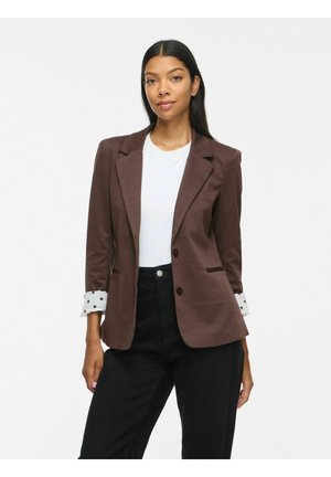 Femme aux longs cheveux foncés porte un blazer marron sur une chemise blanche et un pantalon noir, debout devant un fond blanc uni.