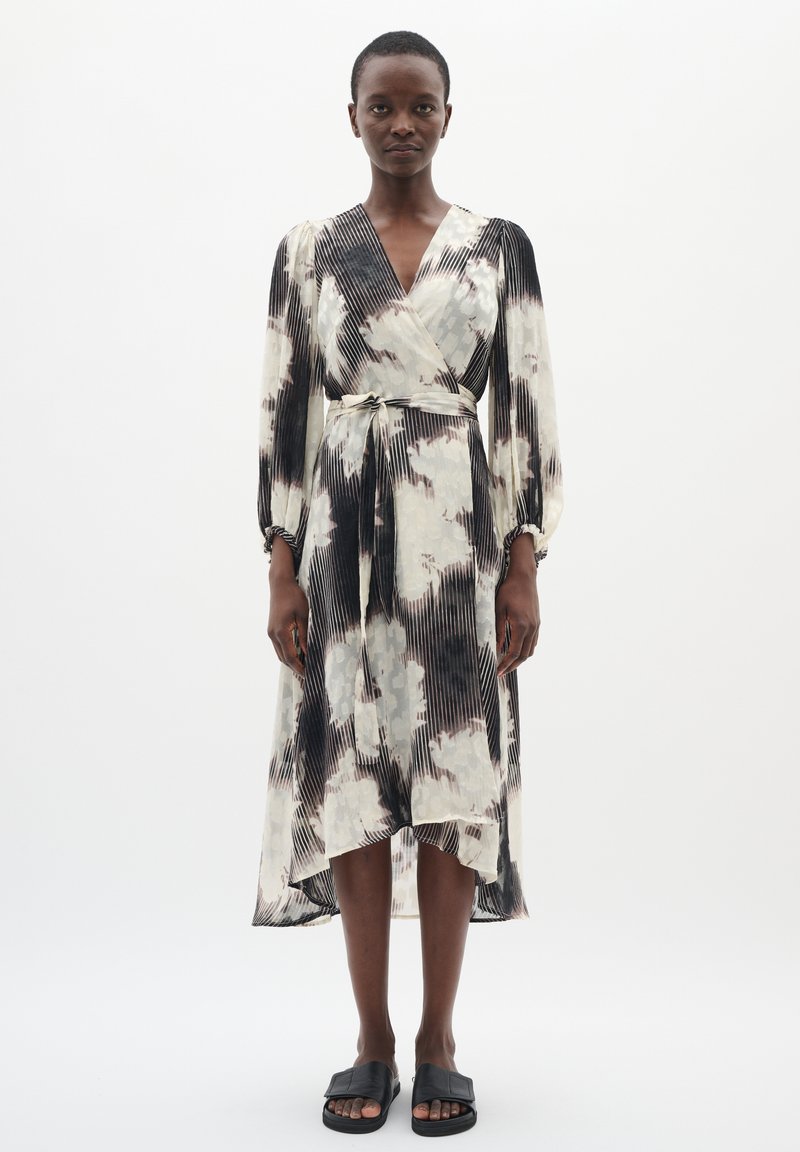 Rochie în pliuri alb-negru, cu un model floral, mâneci lungi și talie legată. Stilizată cu sandale plate negre.