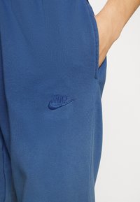 Modré sportovní kalhoty vyrobené z měkkého texturovaného materiálu s viditelným vyšitým logem Nike na bočním kapse.