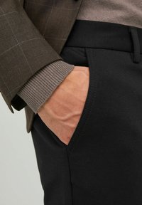 Blazer a quadri marroni con un maglione marrone dalla texture e pantaloni neri. Il primo piano mette in evidenza una mano nella tasca dei pantaloni e i dettagli delle maniche.