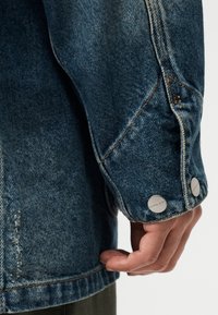 Τζιν μπουφάν σε ξεθωριασμένο μπλε χρώμα, με κοντινή λήψη στο μανίκι. Διακρίνονται ασημένια κουμπιά και ορατές ραφές.