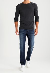 Maglietta grigia scuro a maniche lunghe con maniche raglan, abbinata a jeans in denim blu e sneakers bianche, che mostrano un outfit casual.