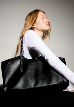 Jeune femme aux longs cheveux portant un haut blanc tient un grand sac cabas en cuir noir sur l'épaule devant un fond uni.