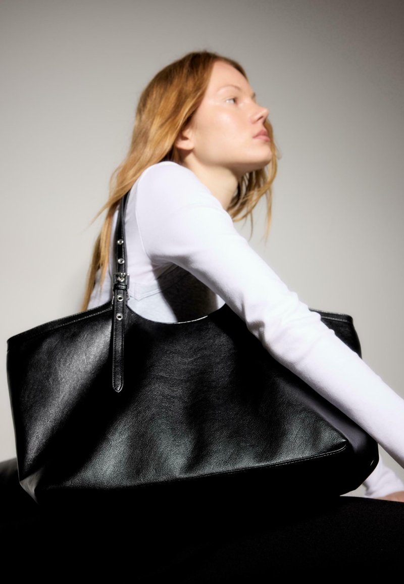 Jeune femme aux longs cheveux portant un haut blanc tient un grand sac cabas en cuir noir sur l'épaule devant un fond uni.