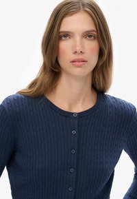 Młoda kobieta z jasnobrązowymi włosami, ubrana w granatowy sweter z dzianiny w warkocze z guzikami, patrzy bezpośrednio w kamerę na białym tle.