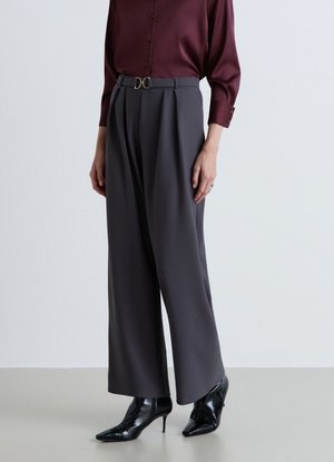 Donna che indossa pantaloni grigi a vita alta e gamba larga con cintura nera, abbinati a una camicia bordeaux con bottoni e stivali neri con tacco a punta.