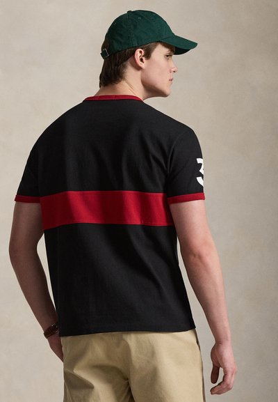 Άνδρας που φοράει μαύρο μπλουζάκι με ευρεία οριζόντια κόκκινη ρίγα, μπεζ παντελόνι και σκουρό πράσινο καπέλο, στραμμένος πλαγίως μπροστά σε απλό φόντο.