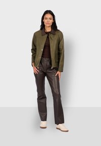 Oliwkowa skórzana kurtka z brązowym kołnierzem, zapinana na zamek, z kieszenią z przodu, zestawiona z ciemnymi skórzanymi spodniami i beżowymi sneakerami.