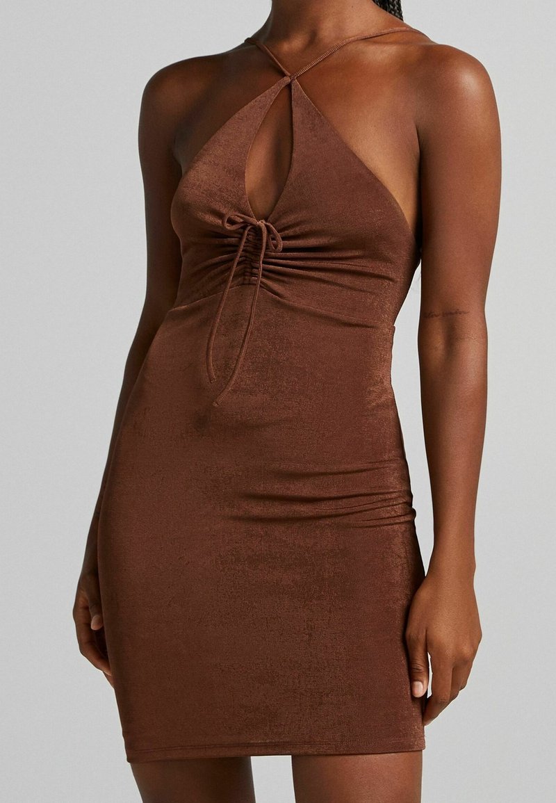 Robe moulante marron sans manches avec col halter, découpe en forme de trou de serrure et détail de nœud à l'avant portée par une personne à la carnation moyenne foncée.