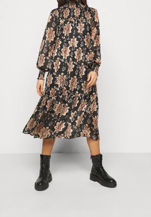 Nainen pukeutunut mustaan midi-mekkoon, jossa on beigenvärisiä kukkakuvioita ja puhvimaiset pitkät hihat, yhdistettynä mustiin nyörikenkiin.