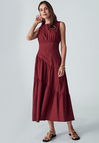 Burgundska brezrokavna obleka s tiered volančki, prilegajočim zgornjim delom in mehkim tekstilom. V kombinaciji s flat sandali, ki imajo zlate poudarke.