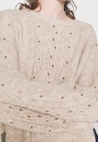 Beige gebreide trui met een gestructureerd patroon, voorzien van gouden accenten en een losse pasvorm. Details omvatten een brede halslijn en geribbelde manchetten.