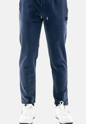 Marineblauwe joggerbroek met elastische boorden en trekkoord in de taille, gedragen met witte sneakers en zwarte sokken op een effen achtergrond.