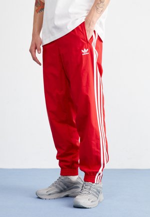Personne portant un pantalon de survêtement rouge Adidas avec des bandes blanches et des baskets grises, debout sur un sol bleu devant un fond blanc.