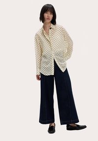 Chemise crème claire à boutons avec des pois noirs, associée à un pantalon en denim foncé à jambes larges. Le modèle se tient devant un fond neutre.
