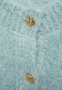Tessuto a maglia testurizzato di colore azzurro chiaro con un aspetto soffice e un dettaglio di bottone rotondo color oro. La vista ravvicinata mostra le fibre e i punti.