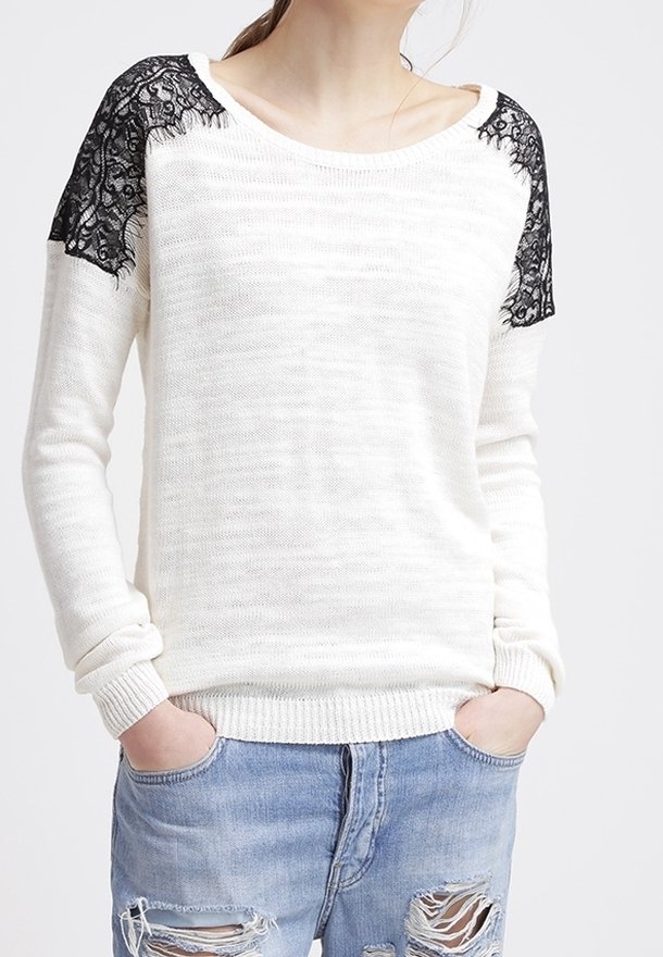 Femme portant un pull en maille blanche avec des panneaux d'épaule en dentelle noire et un jean bleu clair déchiré, les mains dans les poches.