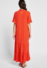 Robe maxi rouge avec de courtes manches volantes, volants superposés au niveau de l'ourlet et un dos en clé. Texture de tissu lisse et léger.