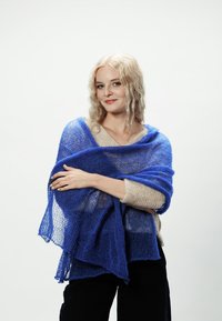 Niebieski dziany szal spoczywa na ramionach, charakteryzując się luźną, przewiewną strukturą. Światło prześwituje, tworząc delikatny efekt gradientu.