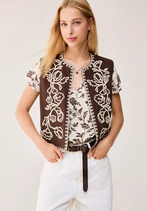 Donna bionda che indossa un gilet marrone ricamato sopra una maglietta floreale, pantaloni bianchi con cintura marrone e gioielli dorati, in piedi con le mani nelle tasche.