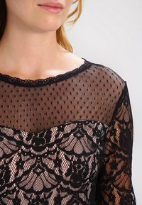 Femme portant un haut en dentelle noire avec une partie supérieure en maille transparente à pois et des manches et un corsage en dentelle florale.