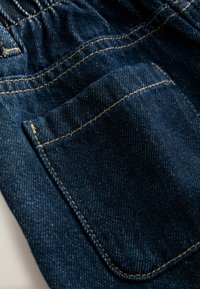 Mørkeblå denimstof med en detaljeret lomme. Har gul syning og en let struktureret overflade. Nærbillede af materialet.