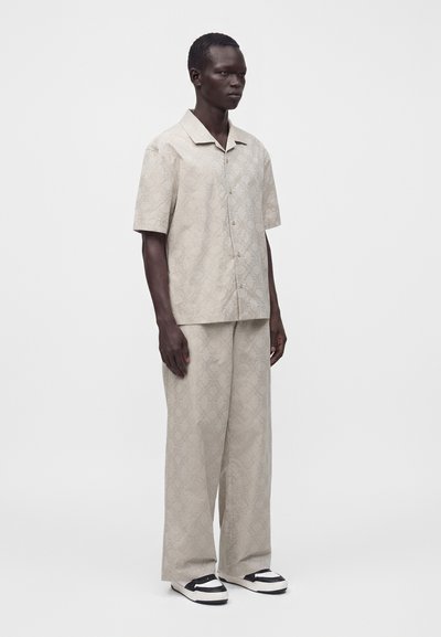 Homme debout portant une chemise beige à manches courtes boutonnée et un pantalon ample assorti avec un motif subtil et des chaussures noires et blanches.