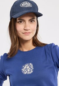 Niebieska bawełniana koszula z krótkimi rękawami, zdobiona białym kwiatowym wzorem na piersi. W komplecie granatowa czapka z kordurowej tkaniny z logo.