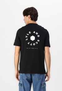 Μαύρο βαμβακερό μπλουζάκι με κοντά μανίκια, με λευκό κείμενο και εξαγωνική γραφιστική στο πίσω μέρος. Χαλαρή εφαρμογή, σε συνδυασμό με μπλε τζιν παντελόνι.