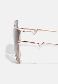Ochelari de soare cu un design geometric. Ramă din metal de culoare roz aurie, lentile gri și accente albe pe brațe, cu un decupaj în formă de zigzag.