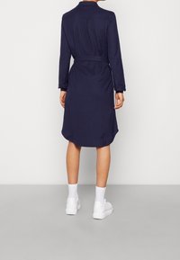 Mörkblå knapp-up klänning med bälte i midjan, långa ärmar och en rundad nederkant. Materialet verkar slätt med en lätt textur.