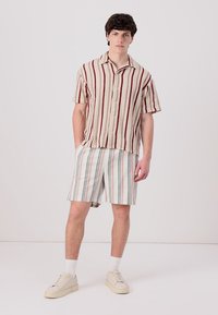 Cămașă scurtă, țesută, cu mânecă scurtă, cu dungi roșii și crem, combinată cu pantaloni scurți cu dungi în culori pastelate. Modelul poartă adidași bej și șosete albe.