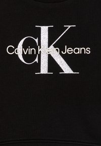 Svart stoff har en metallisk sølvfarge "CK"-logo, med "Calvin Klein Jeans" i hvitt nedenfor. Teksturen er glatt og jevn.