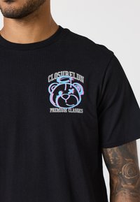 Czarny bawełniany T-shirt z kolorowym graficznym wzorem przedstawiającym niedźwiedzia z aureolą oraz napisem "CLOSURELDN PREMIUM CLASSICS" powyżej.