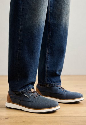 Zapatos de malla azules con acentos de cuero marrón y suelas blancas; cuentan con cordones y una superficie texturizada, combinados con pantalones de mezclilla azul oscuro.
