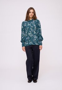 Fließende tealfarbene Bluse mit einem floralen Muster, langen Ärmeln und hohem Kragen, kombiniert mit dunklen Hosen und schwarzen Schuhen.