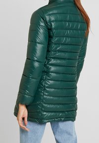 Chaqueta verde, esponjosa, con cuello alto, textura acolchada, acabado brillante y mangas largas. Presenta un logo en el hombro izquierdo.